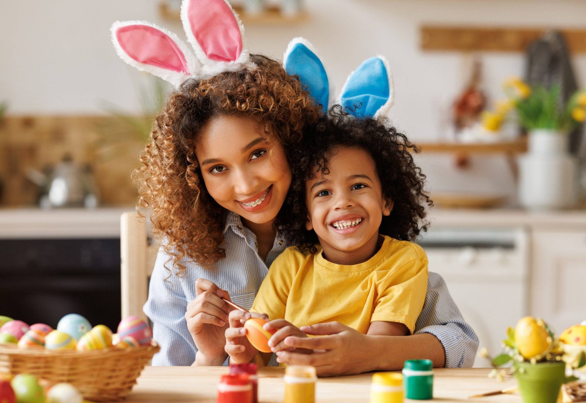 Happy african american family: mother teaching happy little kid soon to decorate Easter eggs while sitting in kitchen Happy african american family: mother teaching happy little kid soon to decorate Easter eggs while sitting in kitchen